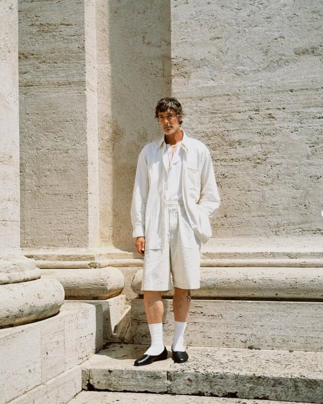 Man in white outfit and white socks wearing black ballet flats against stone wall.