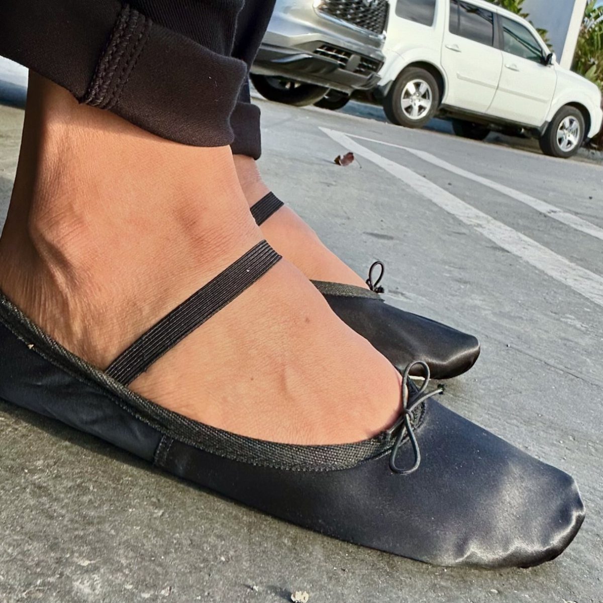 Black satin ballet flats worn with black tailored trousers on a paved street.