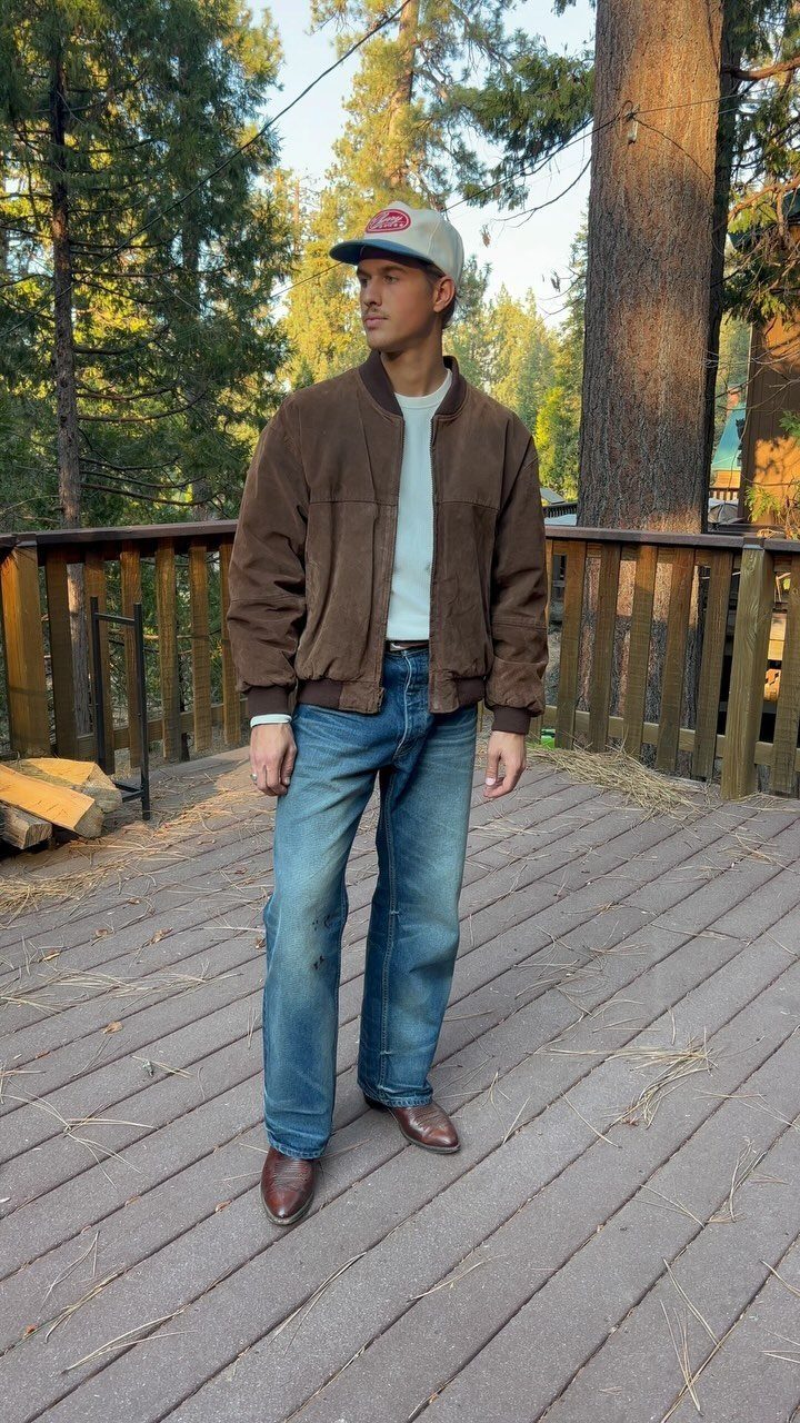 Man in brown jacket, blue jeans, and leather boots standing on a wooden deck.