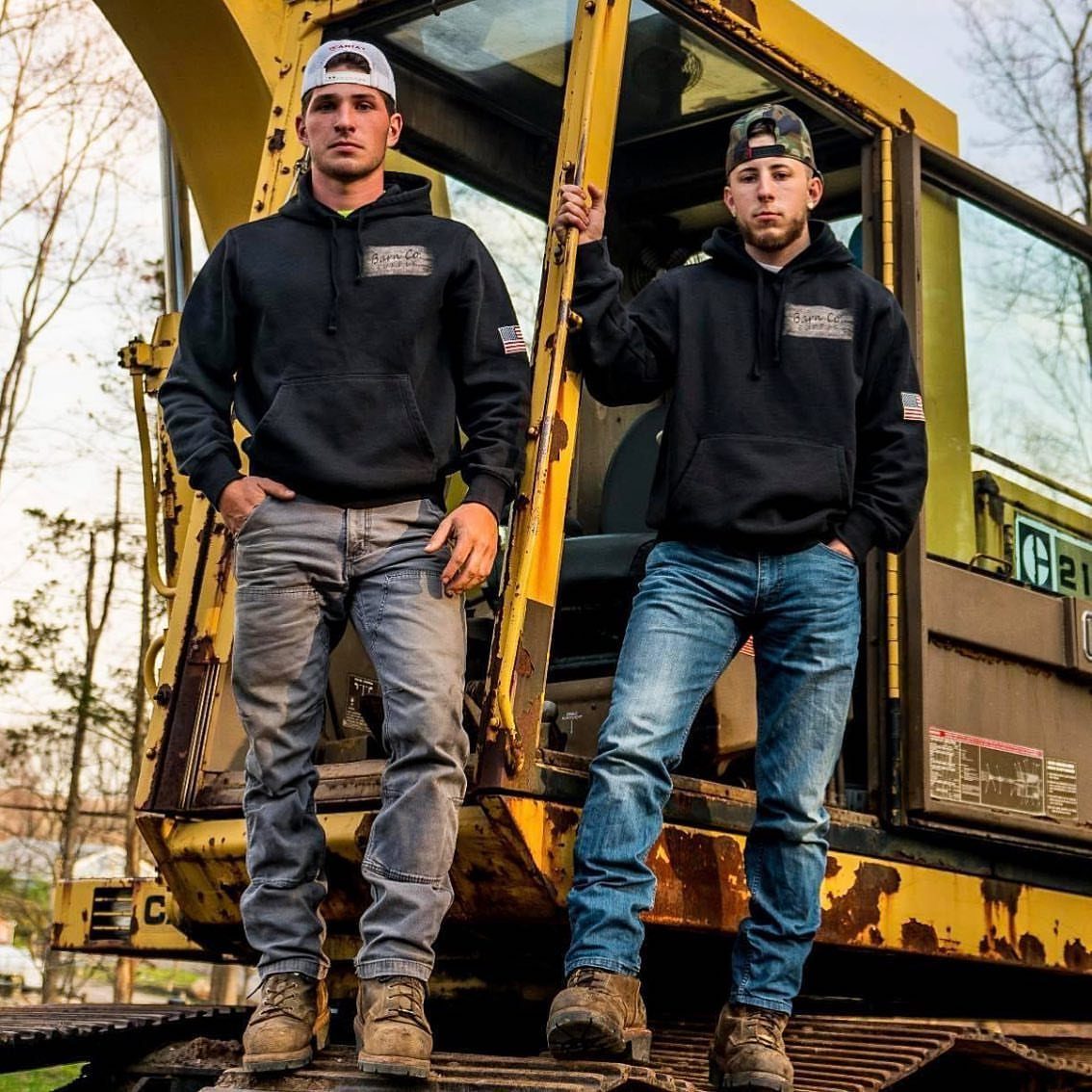 Two men in black hoodies and work boots posing by yellow heavy machinery.
