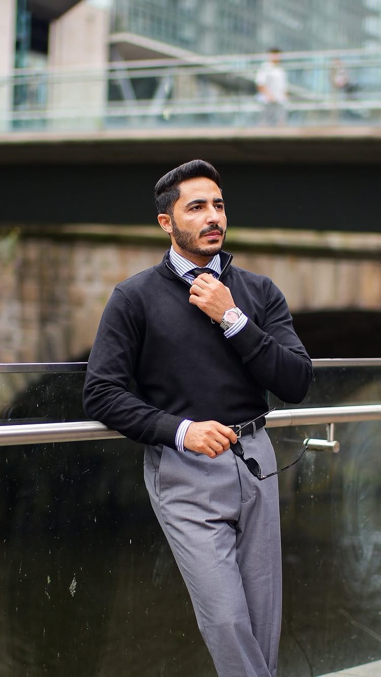 Man in black sweater and gray trousers posing by a city railing.