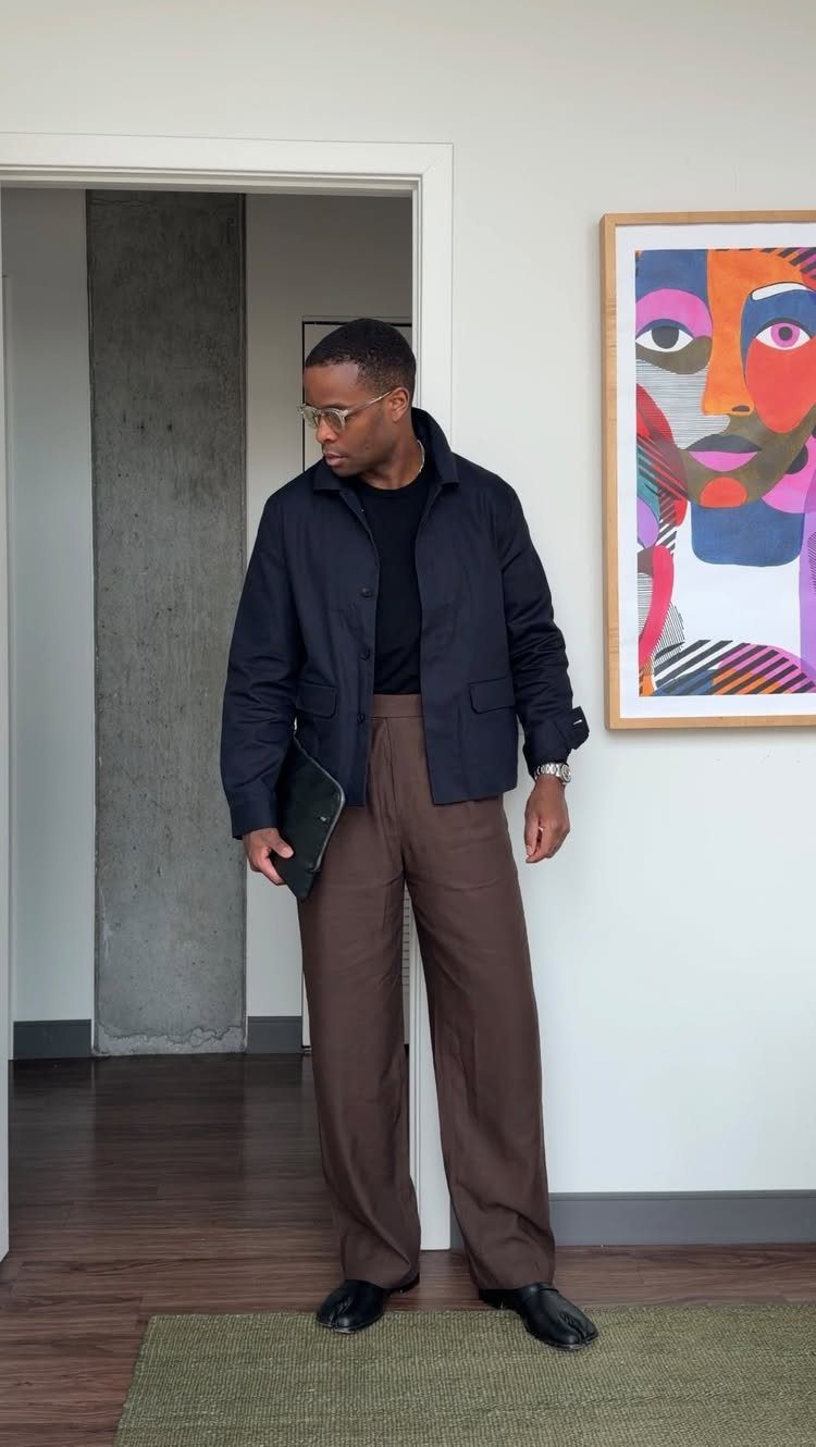 Man wearing a navy blue jacket and brown wide-leg trousers standing indoors.