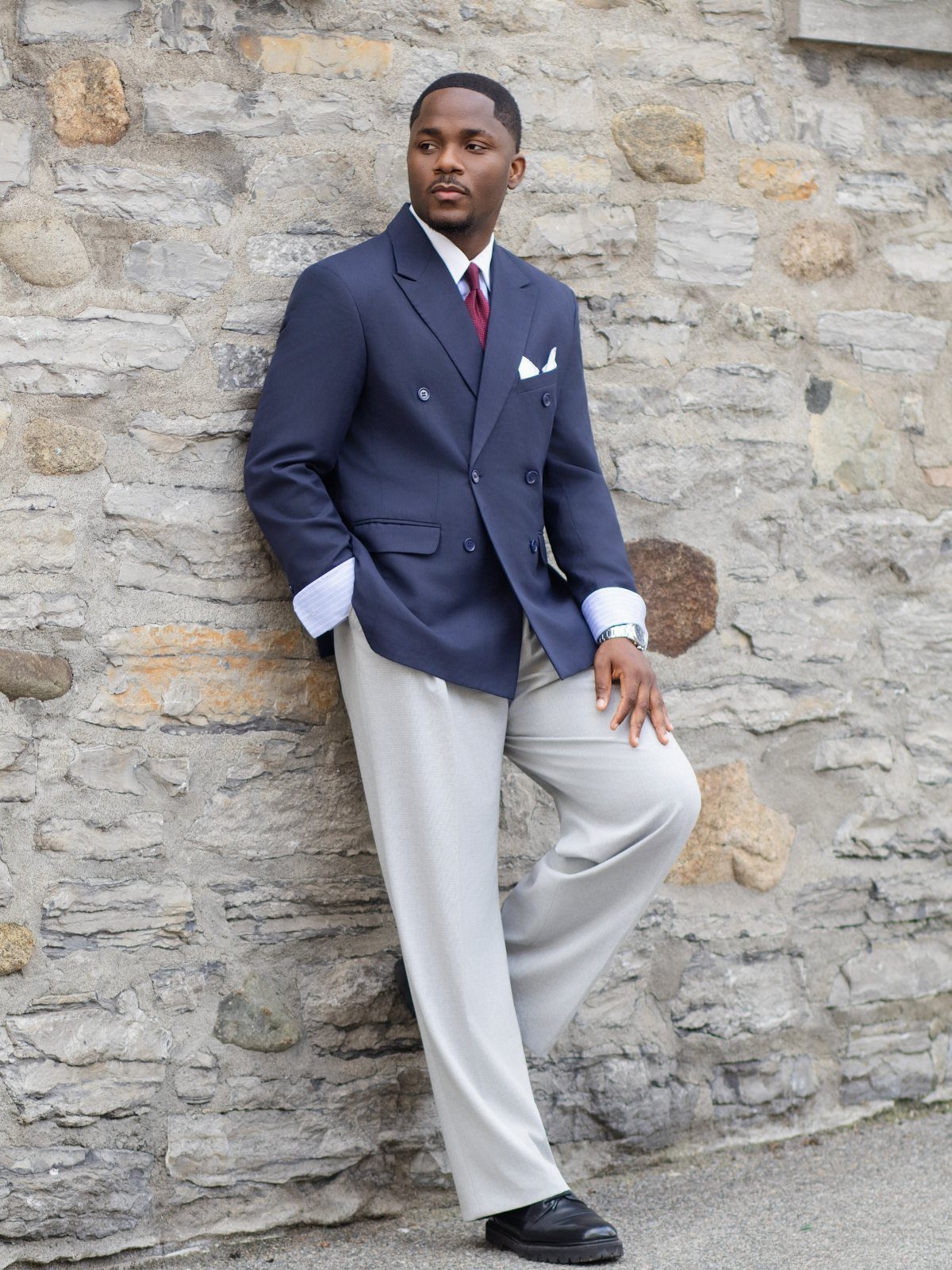 Man in navy blazer and burgundy tie leaning against a stone wall.