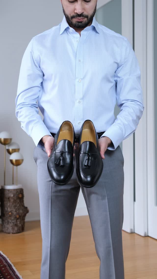 Man in light blue shirt and gray trousers holding black tassel loafers