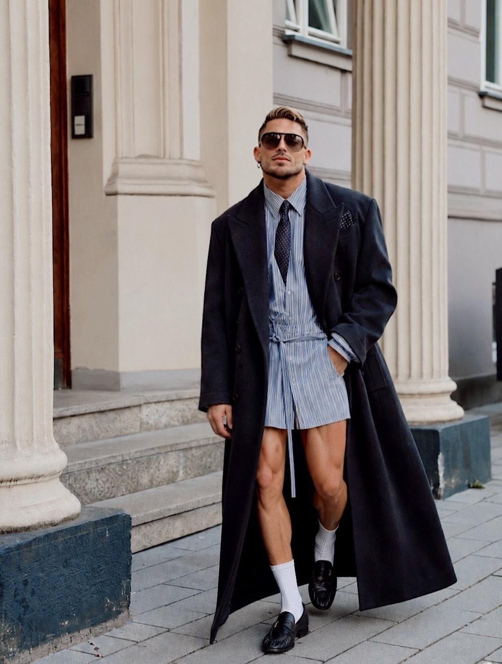 Man in a long black overcoat, striped shirt, and matching shorts walking outdoors.
