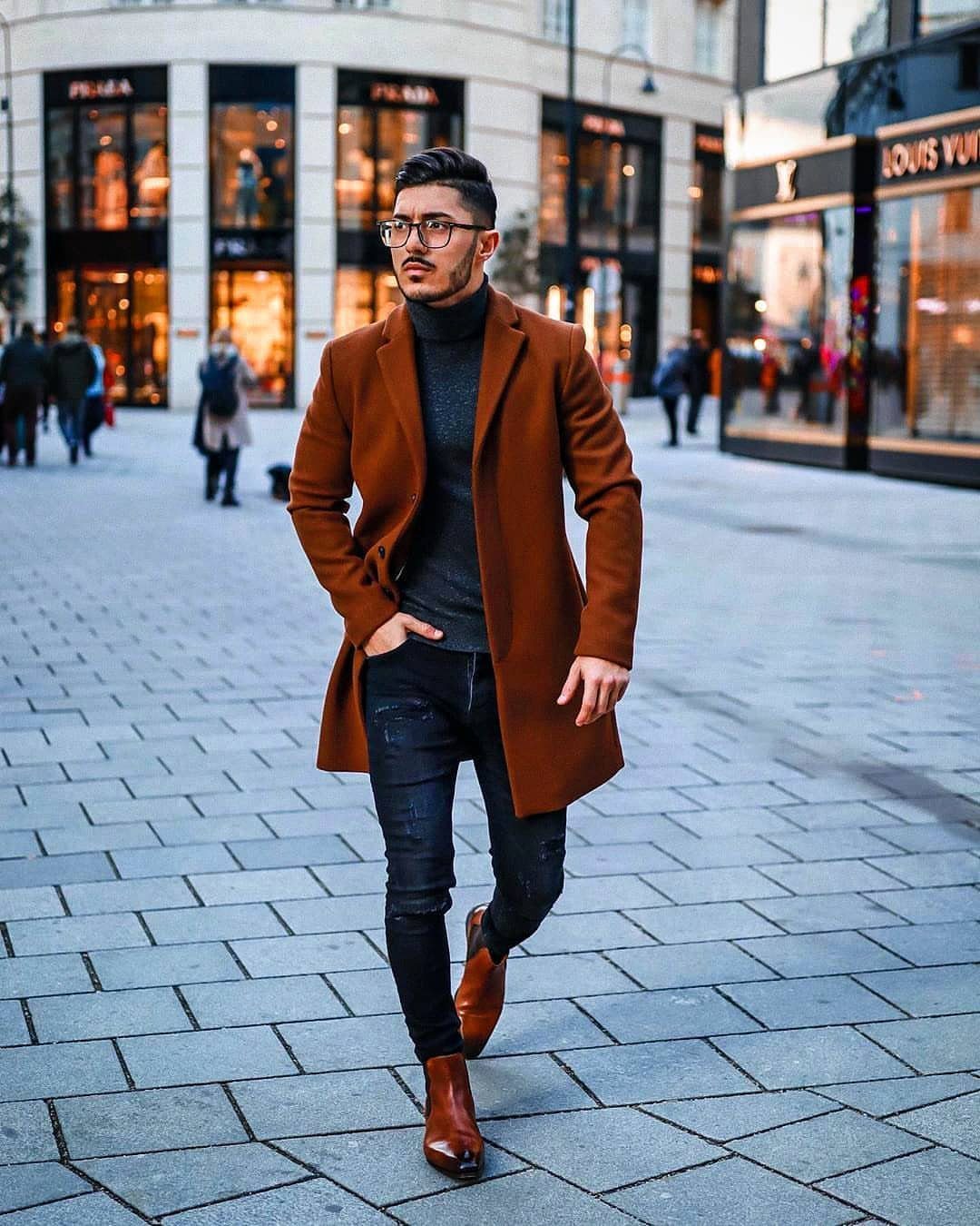 Man in brown overcoat, grey turtleneck, and black jeans on a city street.