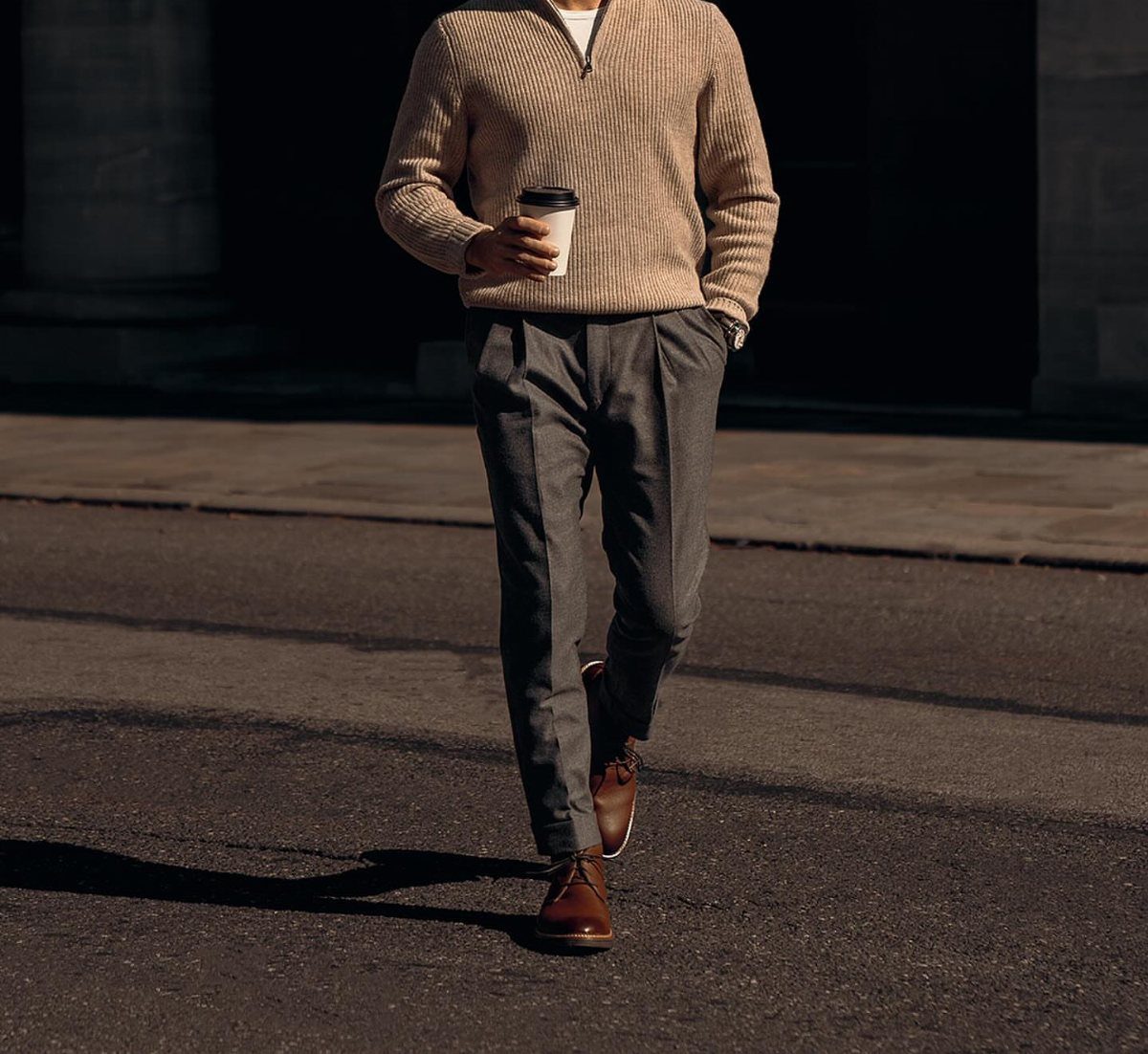 Man in beige ribbed sweater and grey pleated trousers walking on city street.