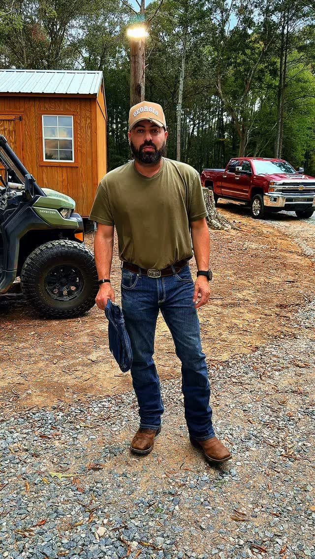 Man wearing olive green t-shirt, blue bootcut jeans, and brown boots outdoors.