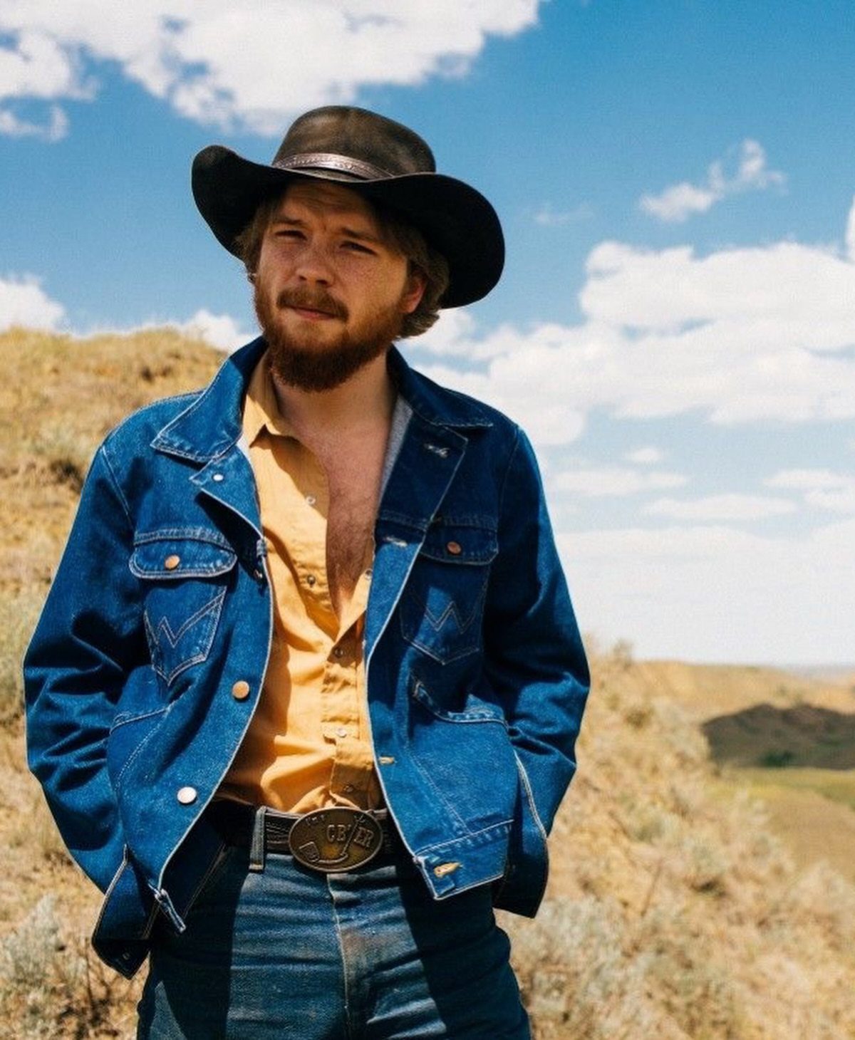 Man in cowboy hat, denim jacket, and yellow shirt posing in a rural landscape