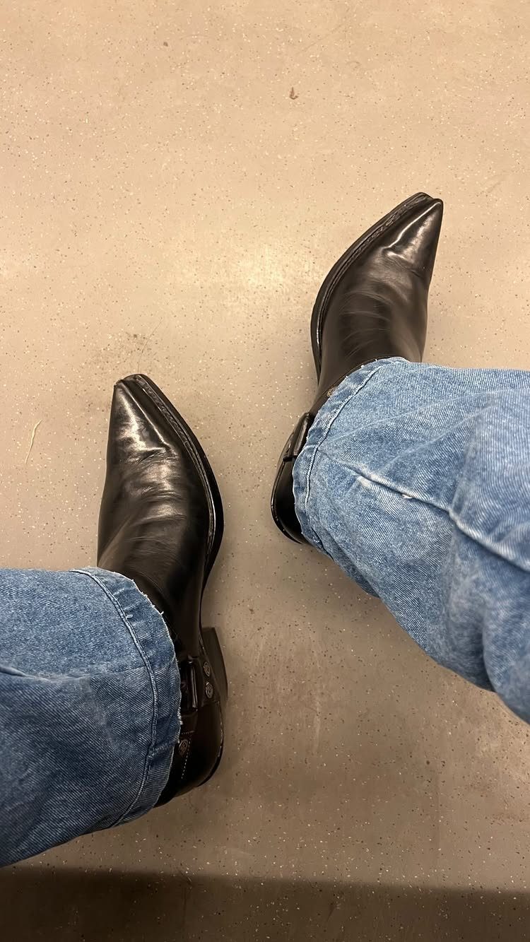 Black leather cowboy boots worn with blue denim jeans on a concrete floor.