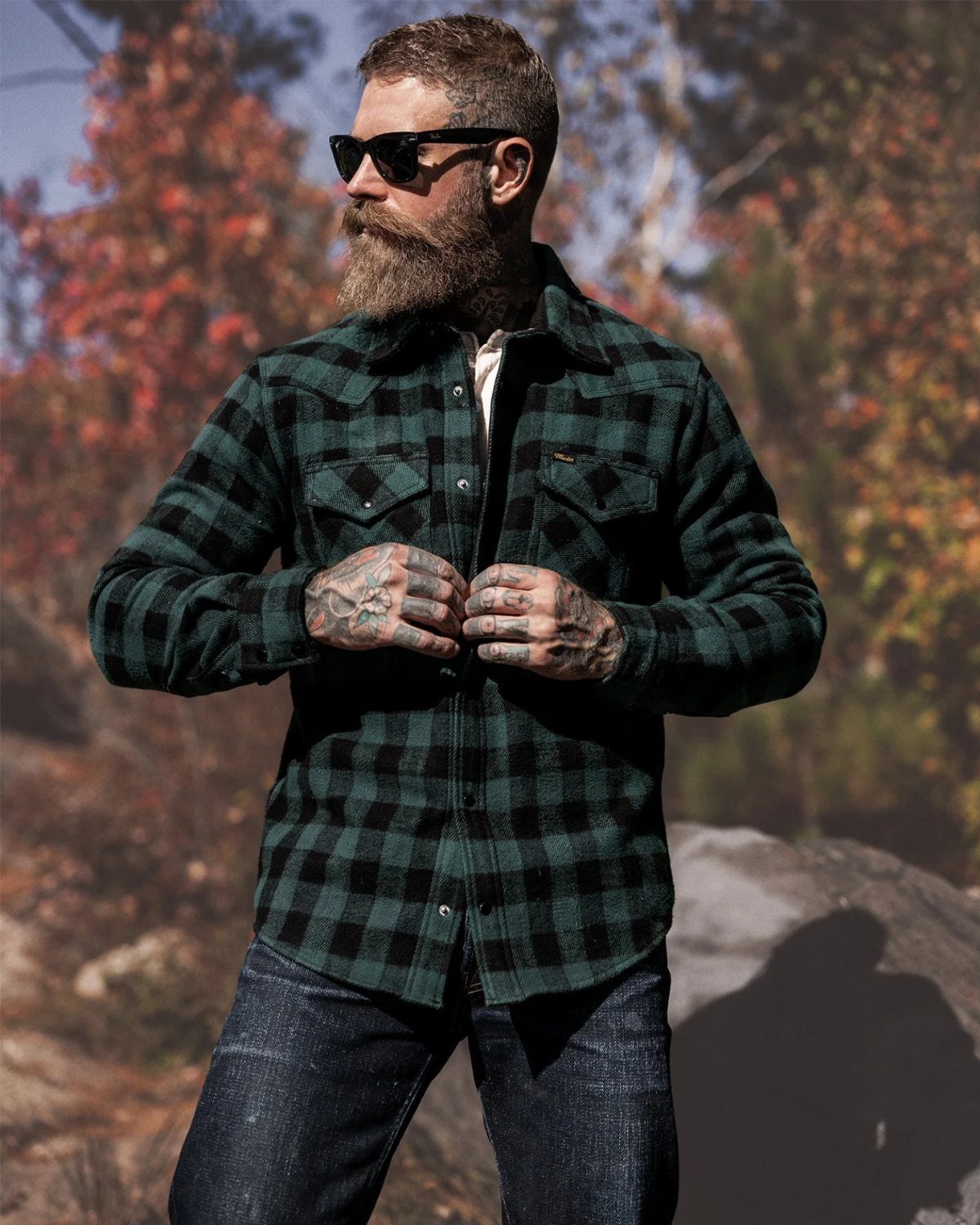 Bearded man wearing a dark green plaid western shirt and dark denim jeans outdoors.