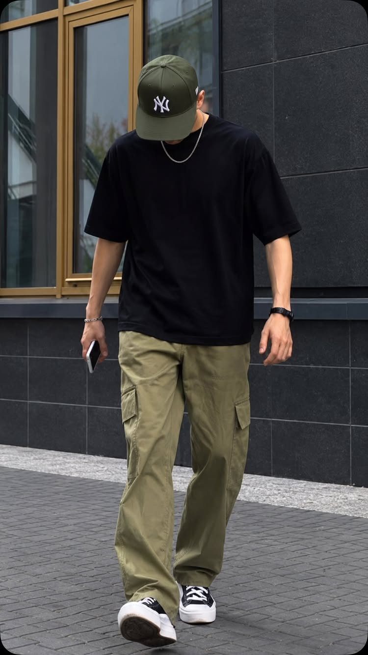 Man in black t-shirt and olive cargo pants walking on a city sidewalk.