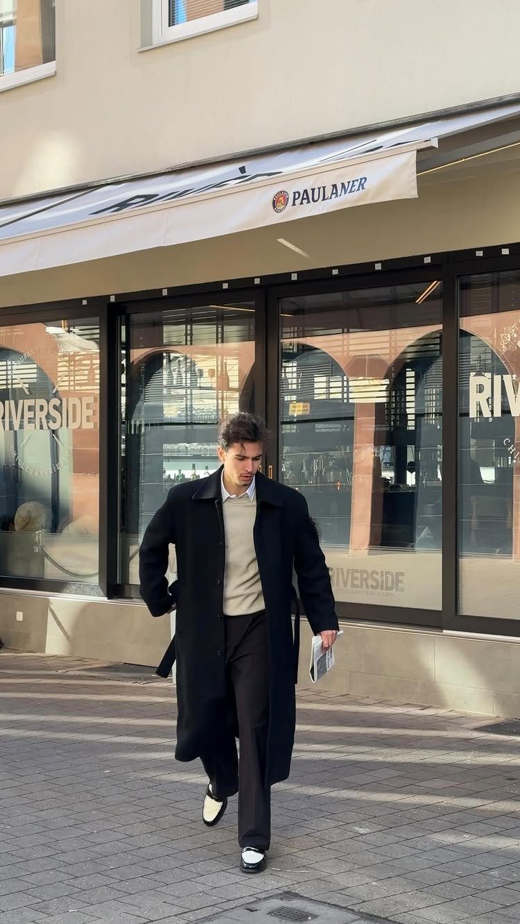 Man wearing a long black overcoat and beige sweater walking on a city street