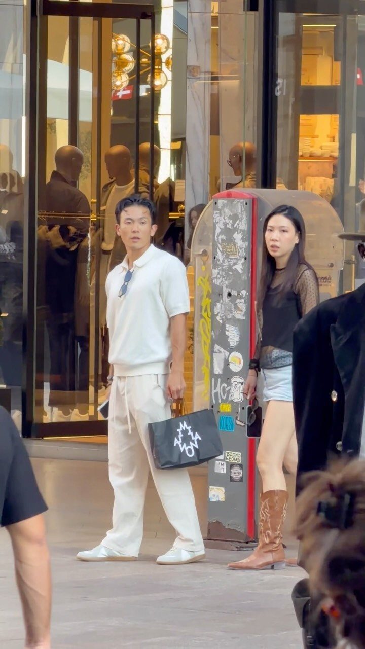 Man in all-white polo and wide-leg trousers holding a black handbag on city street.