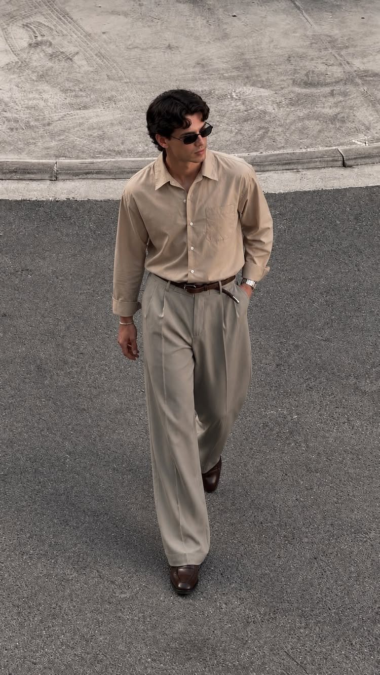 Man wearing a tan shirt and light grey wide-leg trousers walking on asphalt.