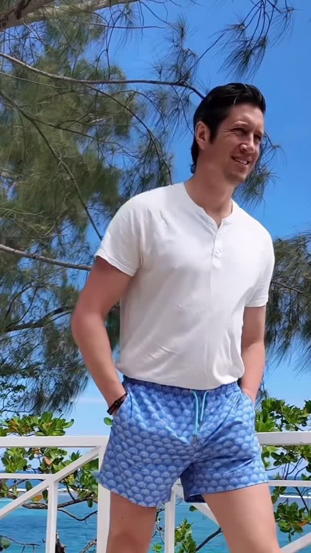 Man wearing a white henley shirt and blue patterned swim shorts on a balcony.