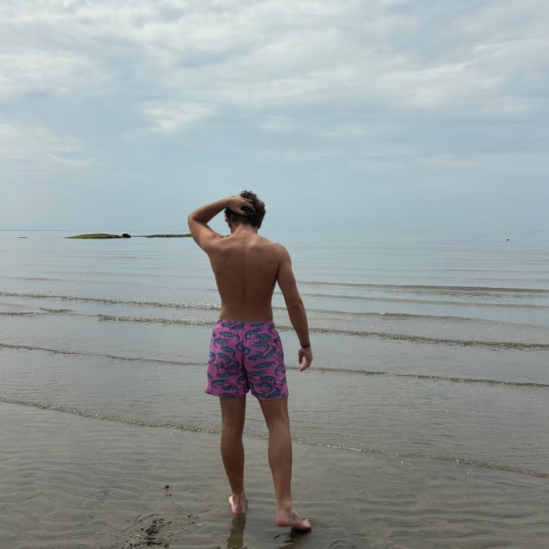 Man standing on a beach wearing pink and blue patterned swim trunks.