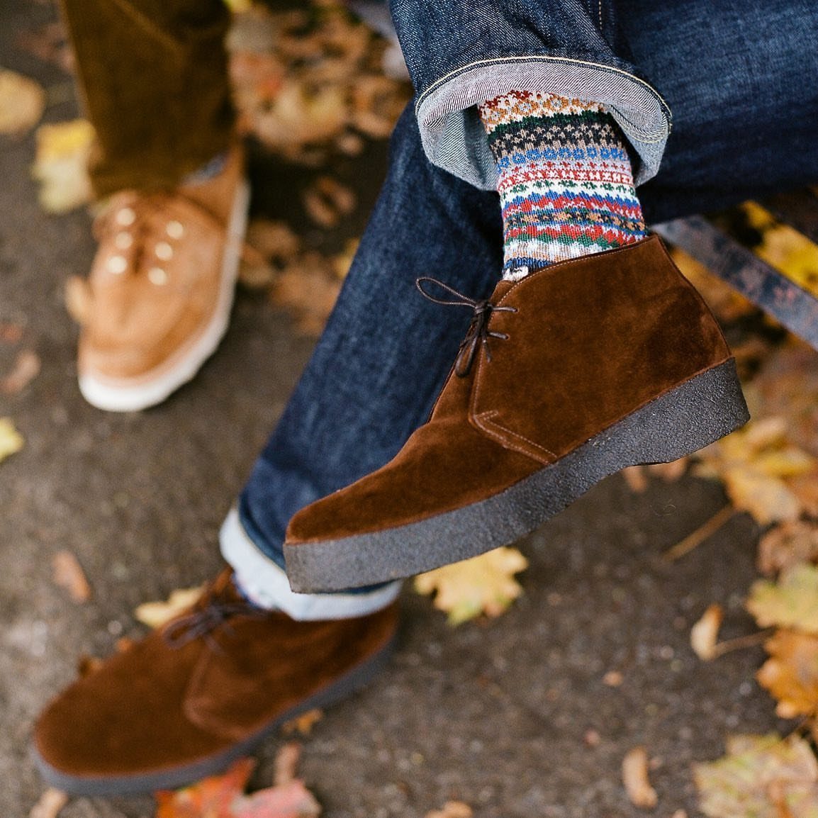 Brown suede chukka boots with colorful patterned socks and cuffed blue jeans.
