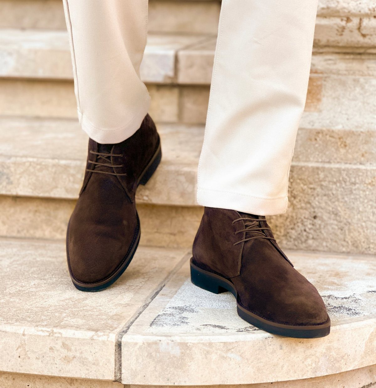 Dark brown suede chukka boots worn with off-white trousers on stone steps.