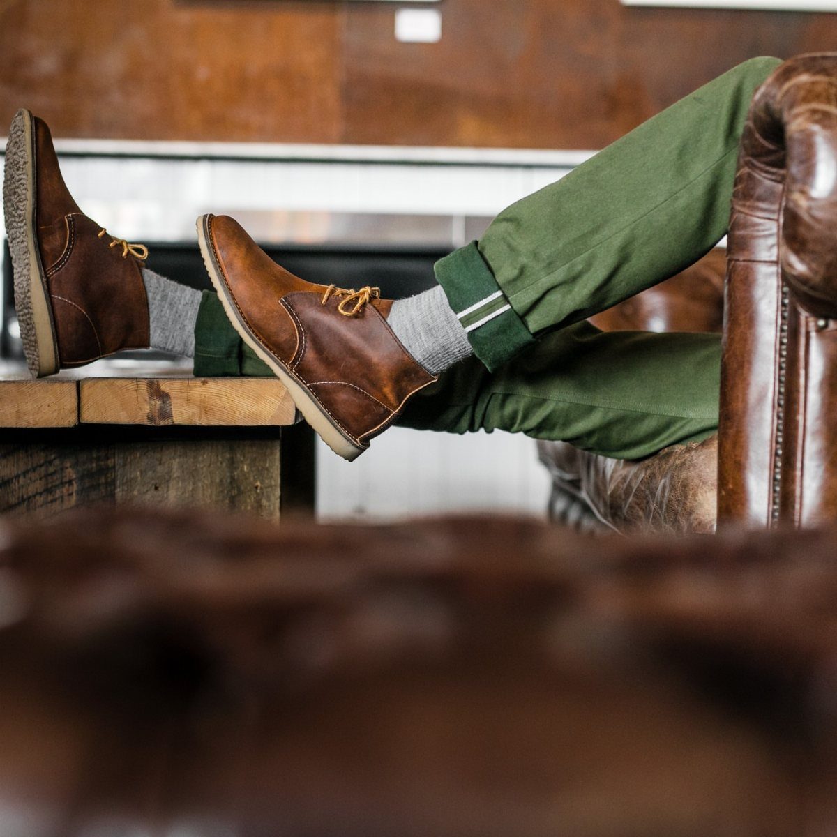Brown leather chukka boots worn with green trousers and grey socks resting on a table.
