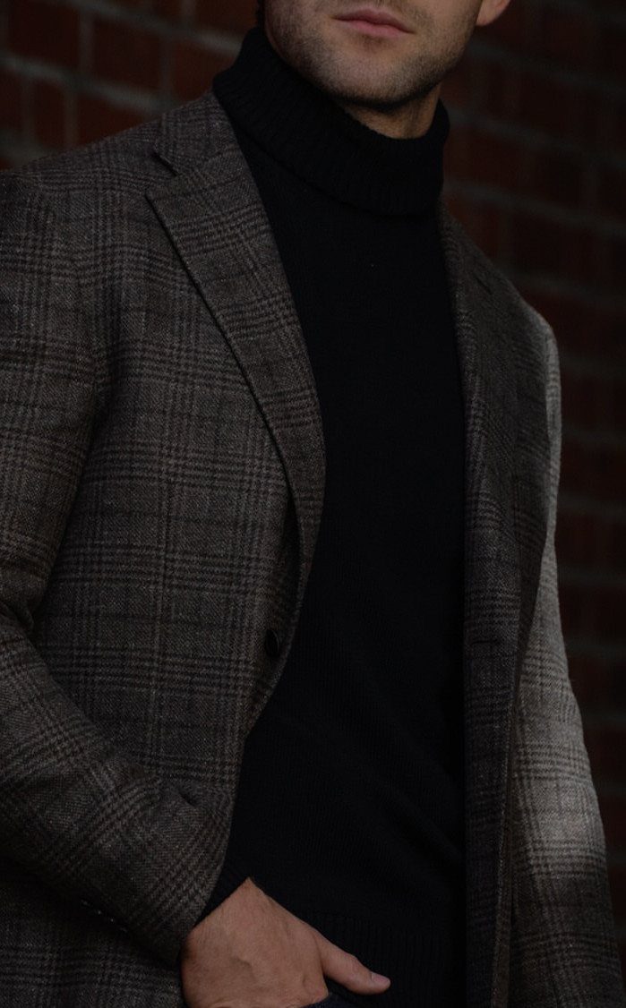 Man wearing a brown plaid tailored blazer and black turtleneck against brick wall