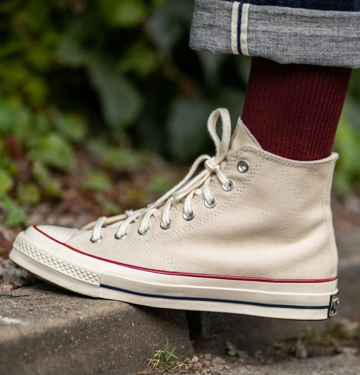 Cream high-top Converse shoe with maroon socks and cuffed grey trousers.