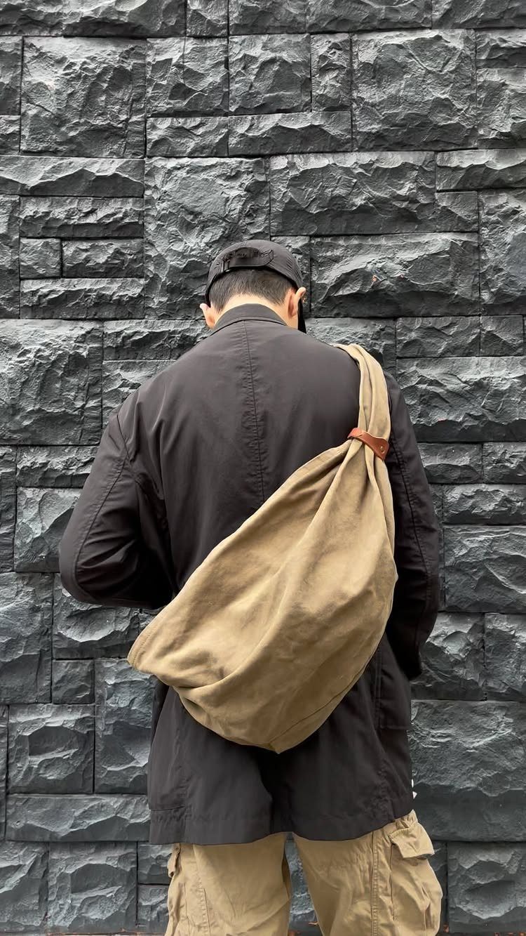 Men's Fashion Crossbody Bag: 6 Styles Worth Carrying 3 Man wearing a tan heavy canvas crossbody bag and black jacket against stone wall