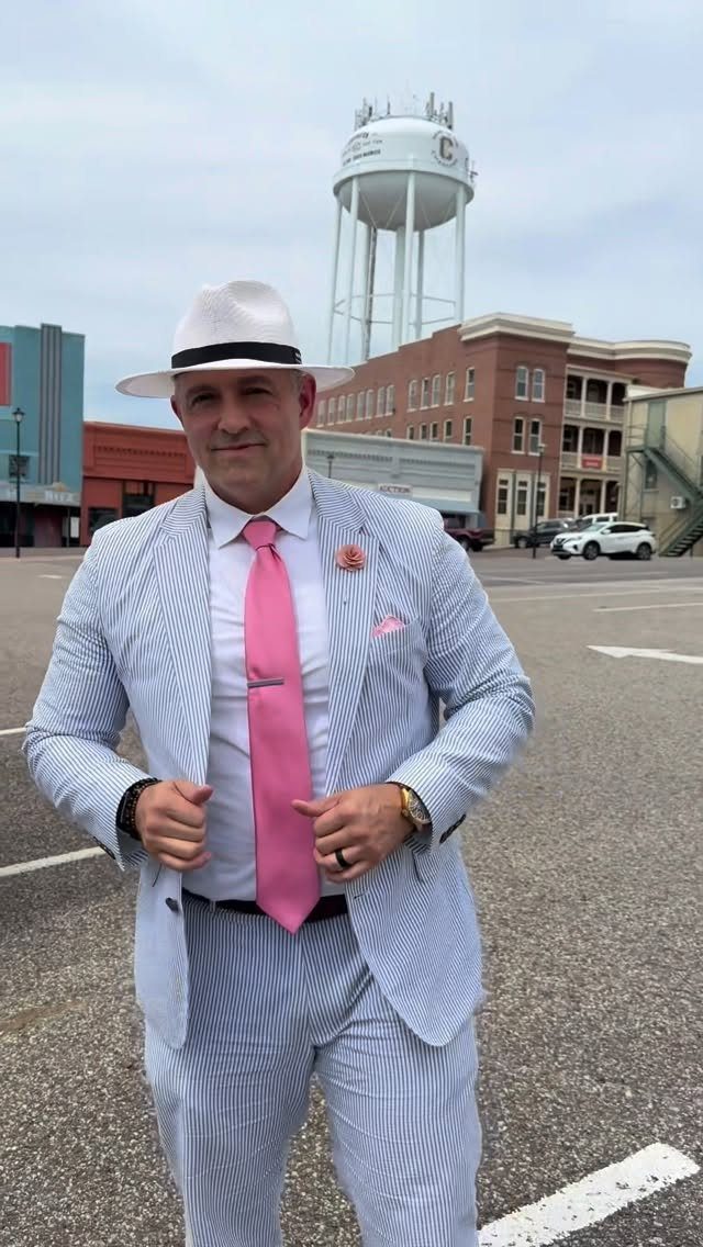 Man in seersucker suit, pink tie, and white fedora standing in a parking lot.