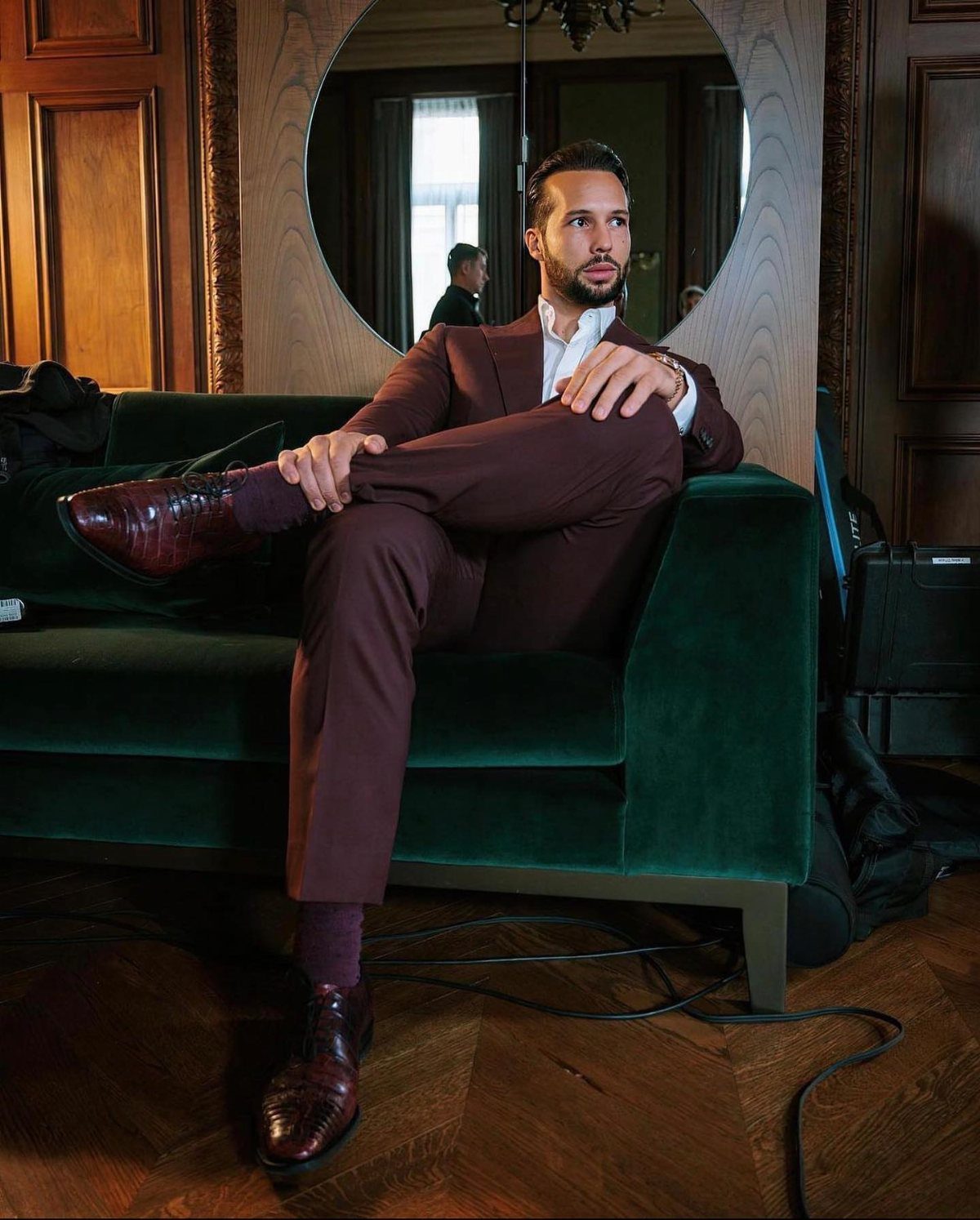 Man in a deep burgundy suit sitting on a green velvet sofa.