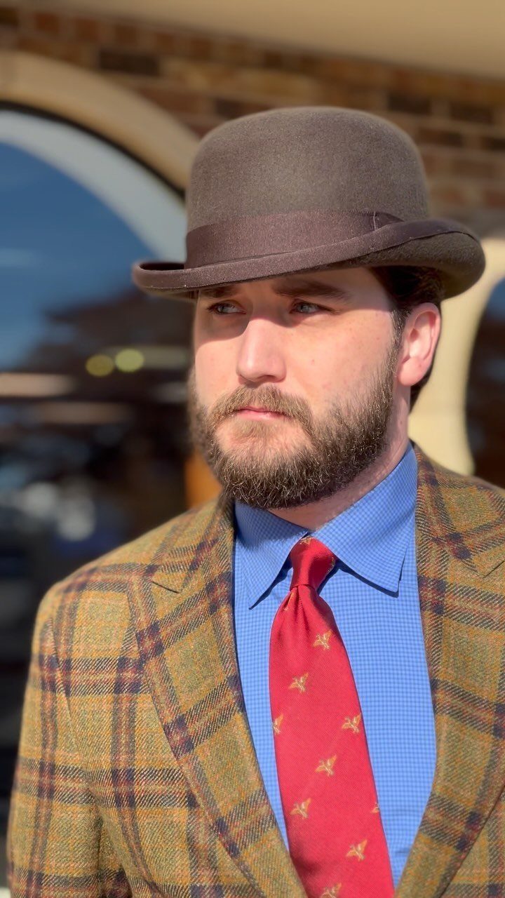 Man wearing a brown bowler hat, plaid blazer, blue shirt, and red tie.