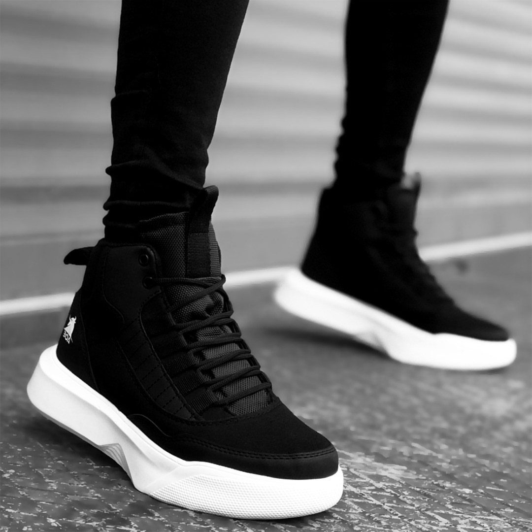 Black high-top sneakers with white soles worn with black pants on grey ground.