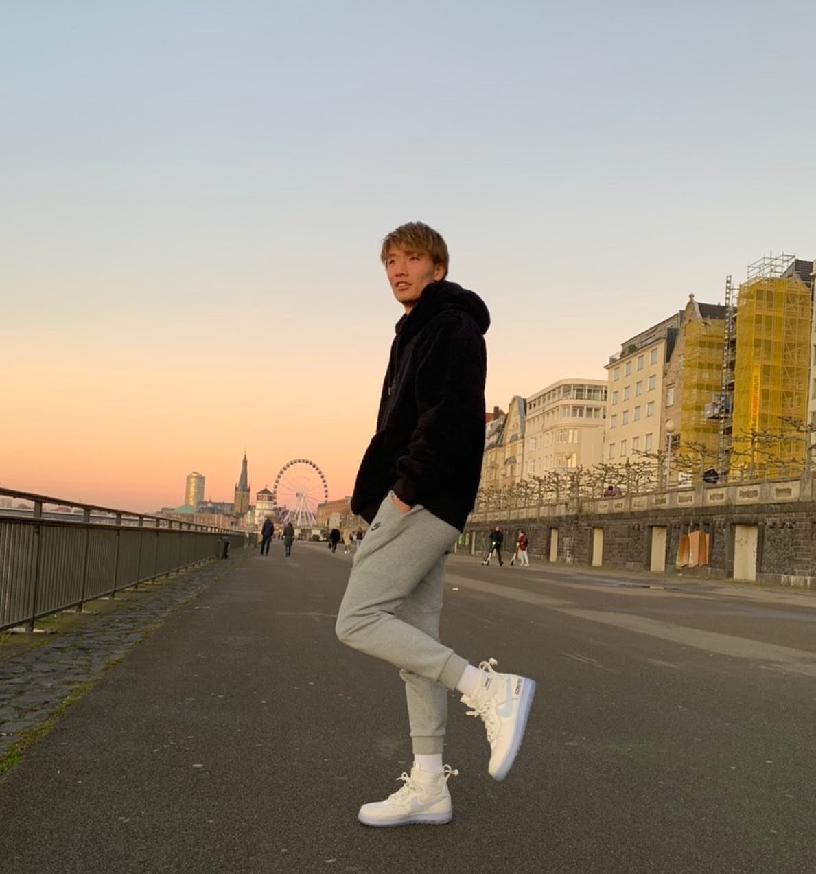 Man in black hoodie and grey joggers wearing white high-top sneakers outdoors.