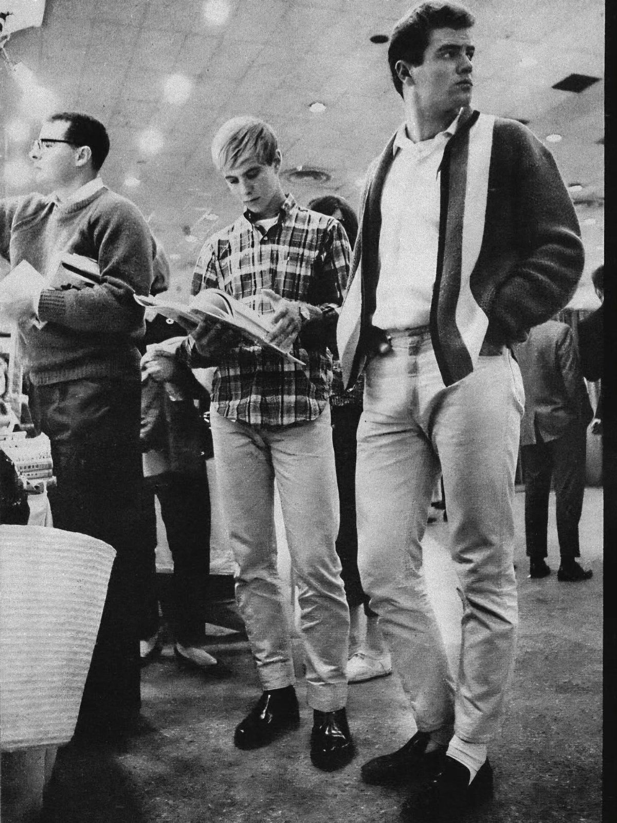 Three young men wearing 1960s style cardigans, plaid shirts, and trousers in a store.