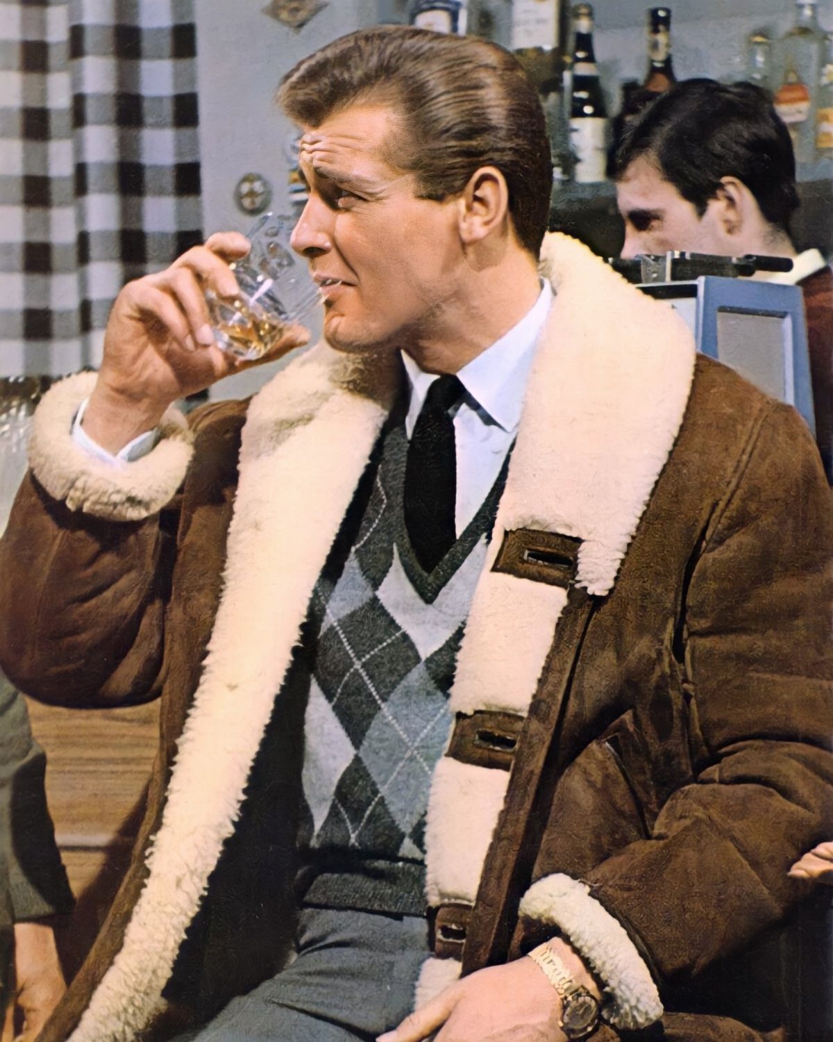 Man in 1960s fashion wearing a shearling coat and argyle sweater drinking whiskey.