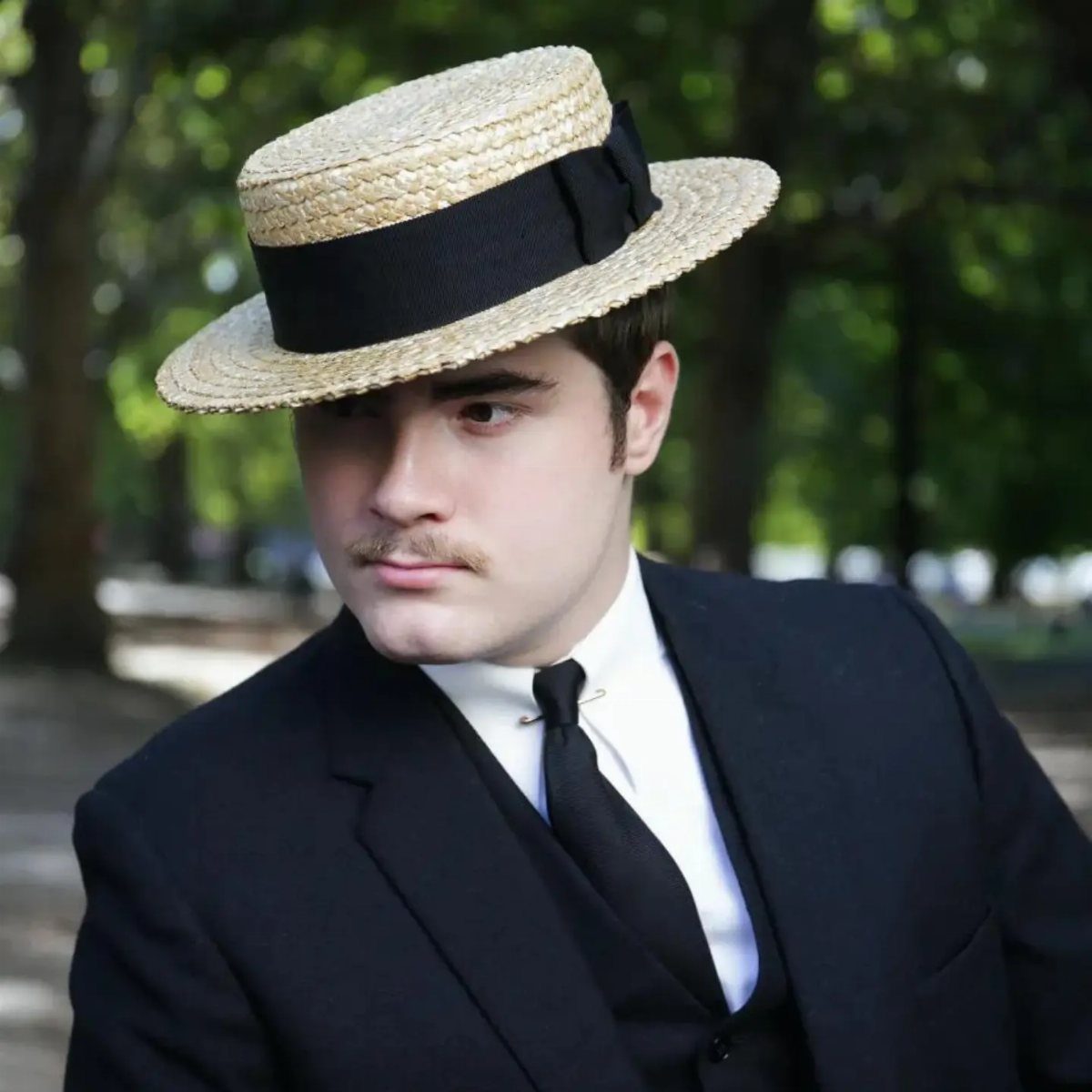 Man in a vintage dark suit and straw boater hat in a park.