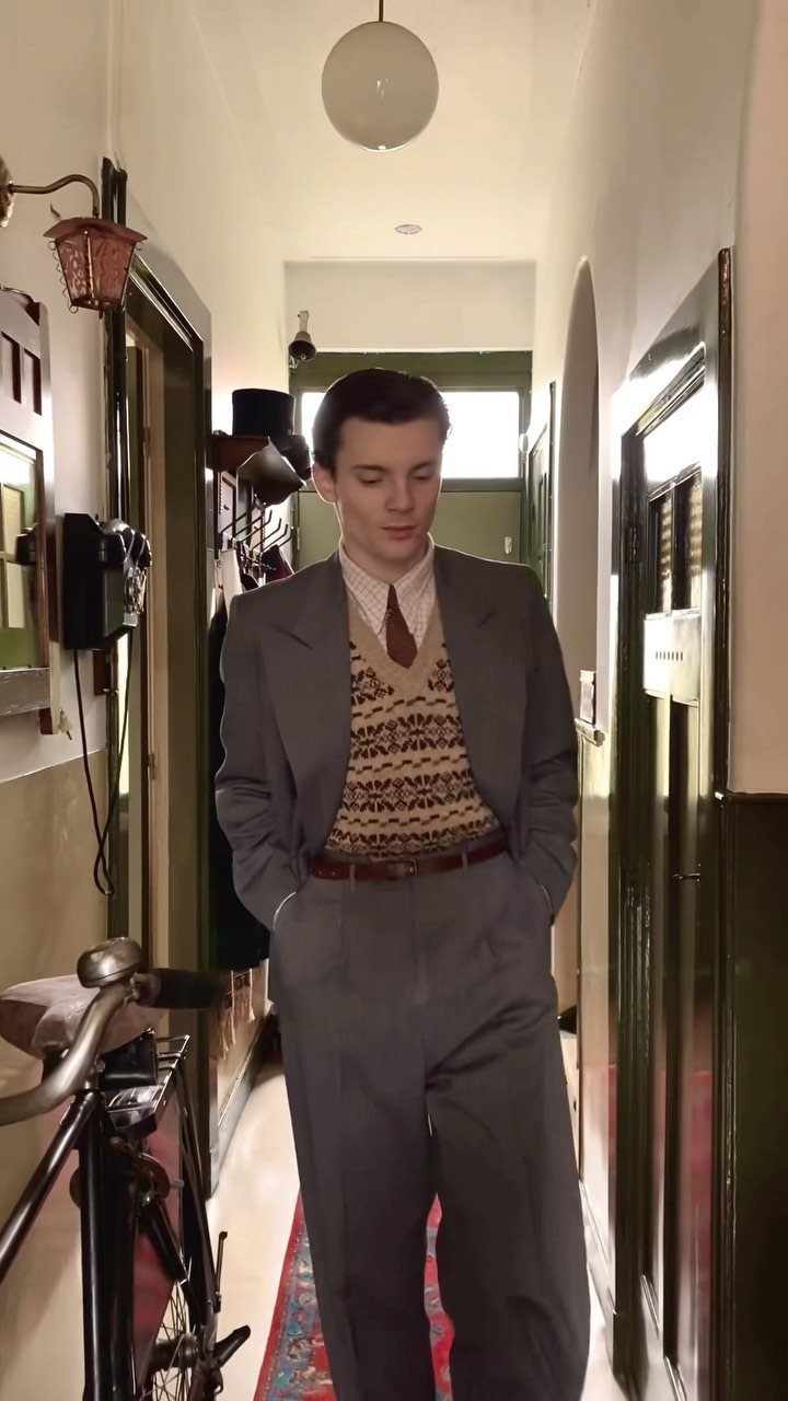 Man in a grey suit and patterned sweater vest standing in a hallway.