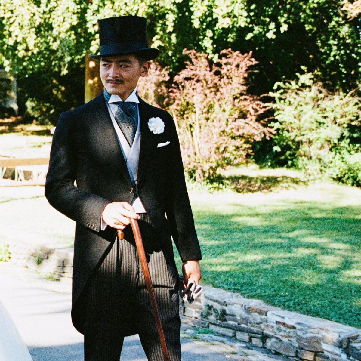 Man in a black top hat and morning coat holding a wooden cane.