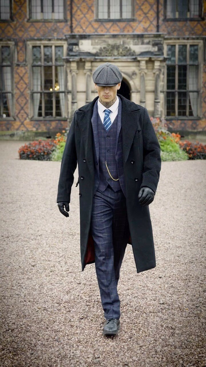Man in a vintage 1920s style purple plaid suit, black overcoat, and flat cap.