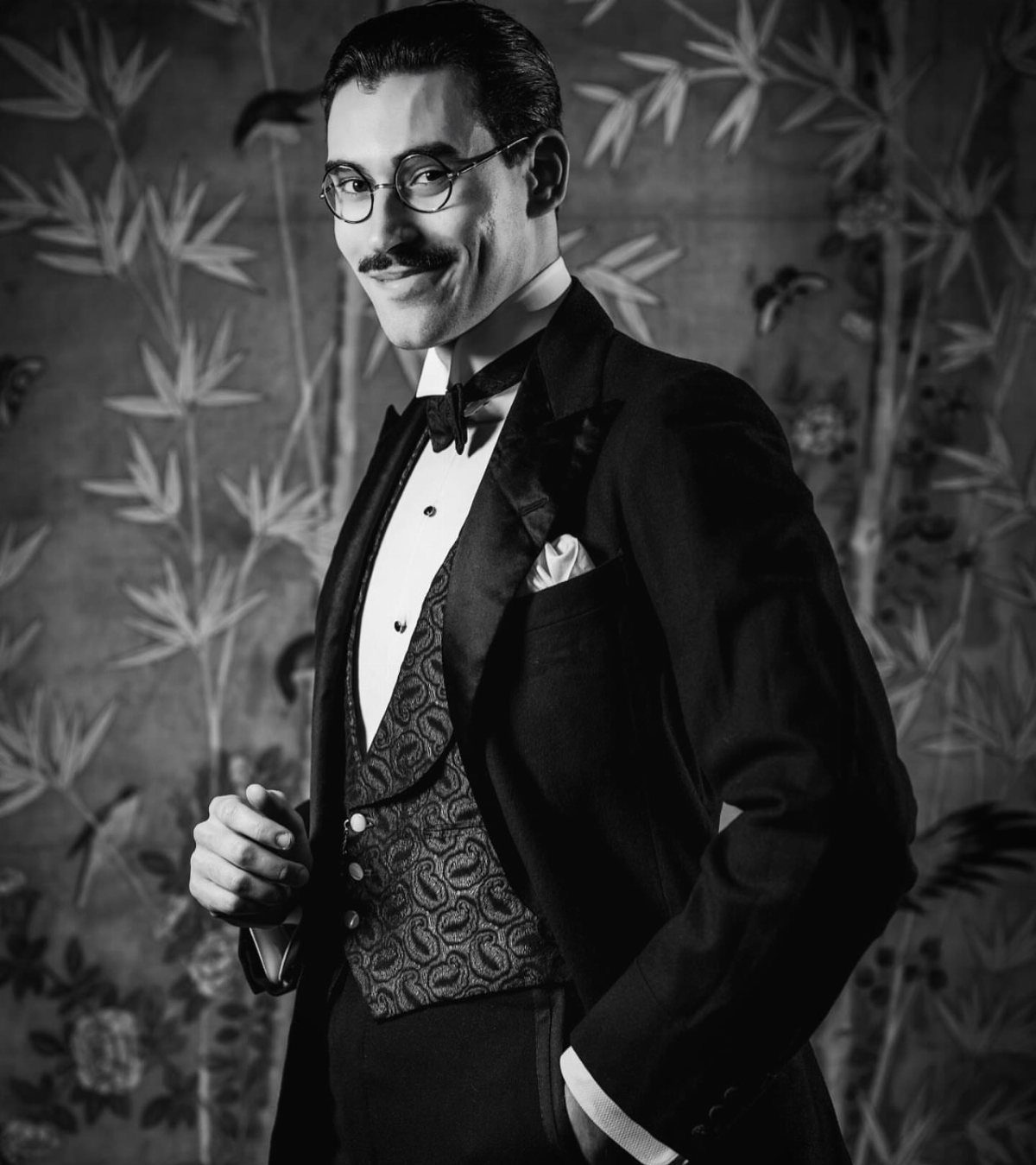 Man in 1920s tuxedo with patterned waistcoat and round glasses posing against bamboo background.