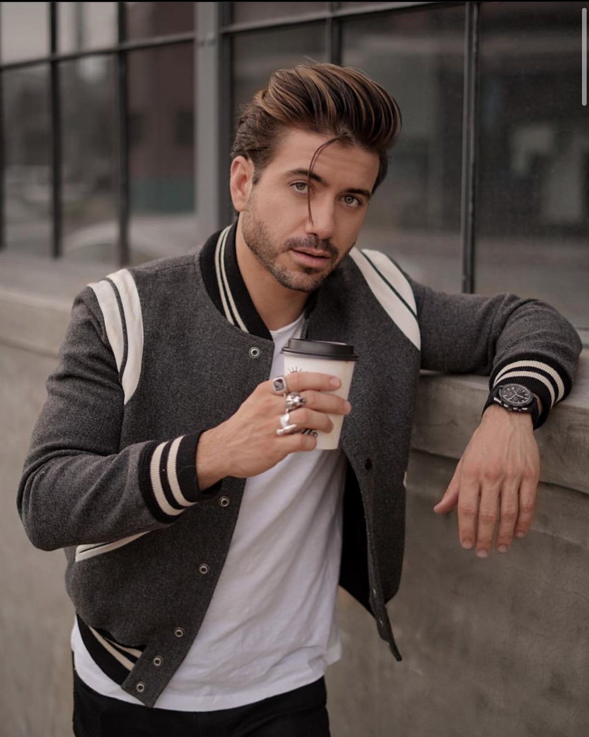 Man in a grey varsity jacket holding a coffee cup in an urban setting.