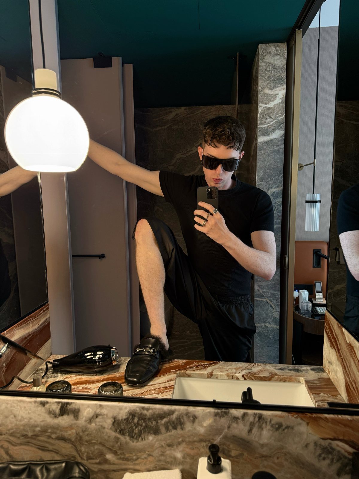 Man in all-black outfit and sunglasses taking a mirror selfie in a bathroom.