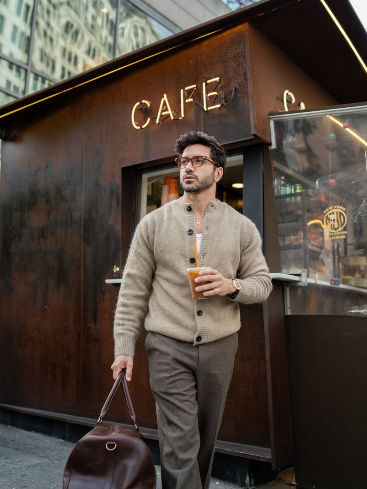 Man in beige cardigan and brown trousers posing outside a modern cafe.