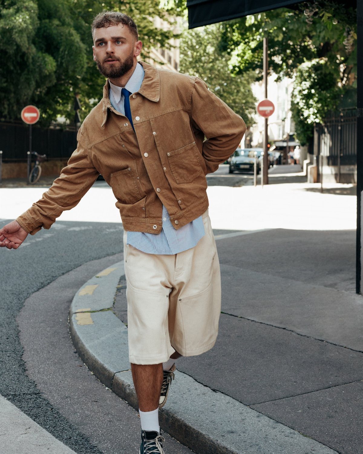 Man wearing a brown suede jacket and cream shorts walking on a city street.