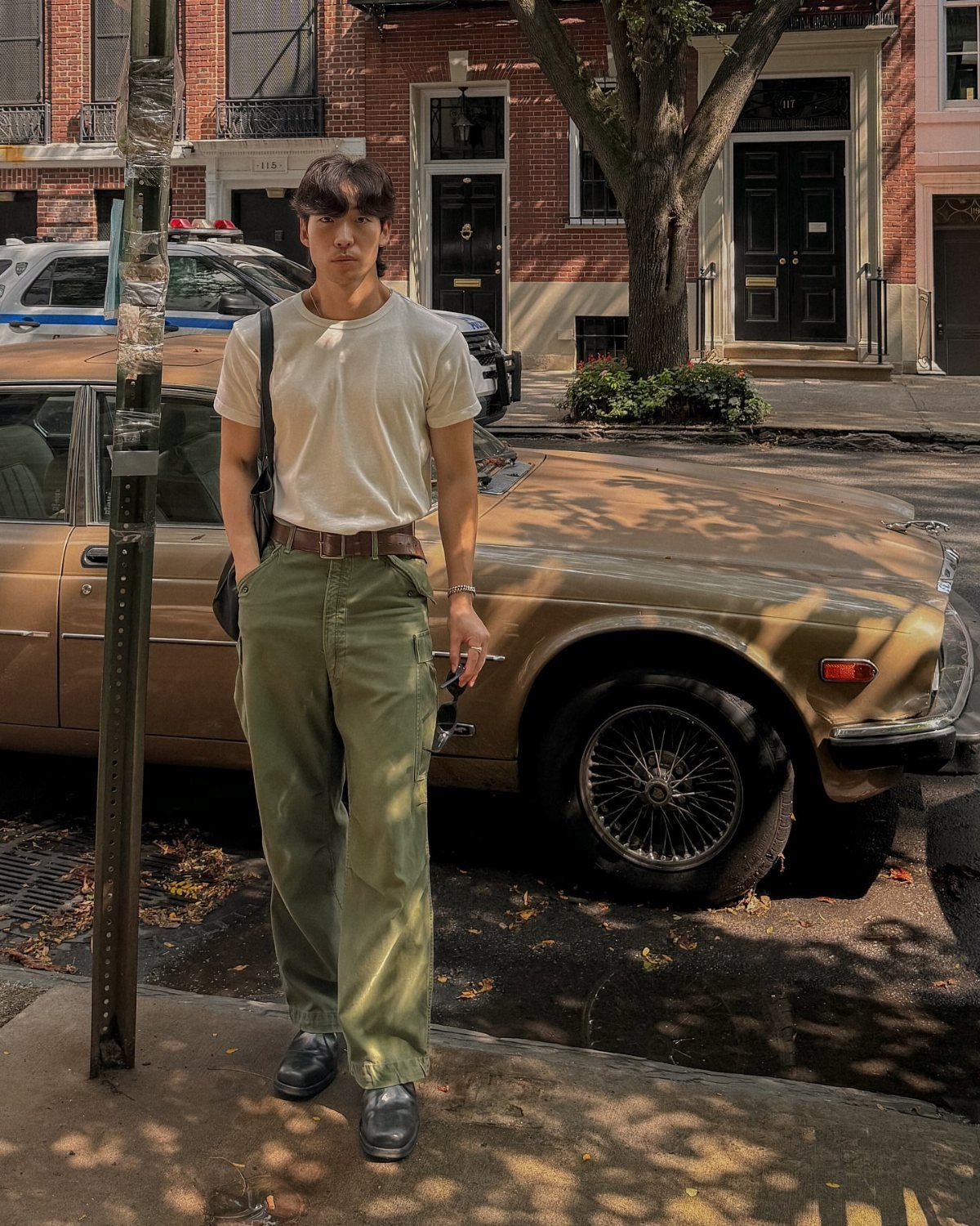 Man in white t-shirt and olive trousers posing by a vintage gold car.