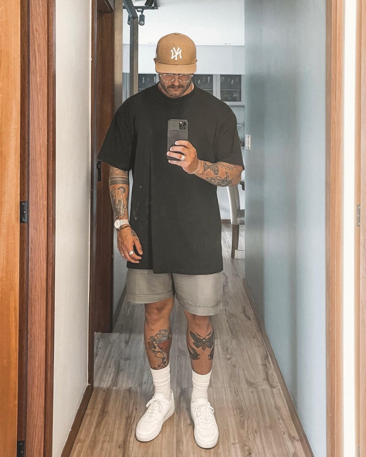 Man in oversized black t-shirt, grey shorts, and tan cap taking mirror selfie
