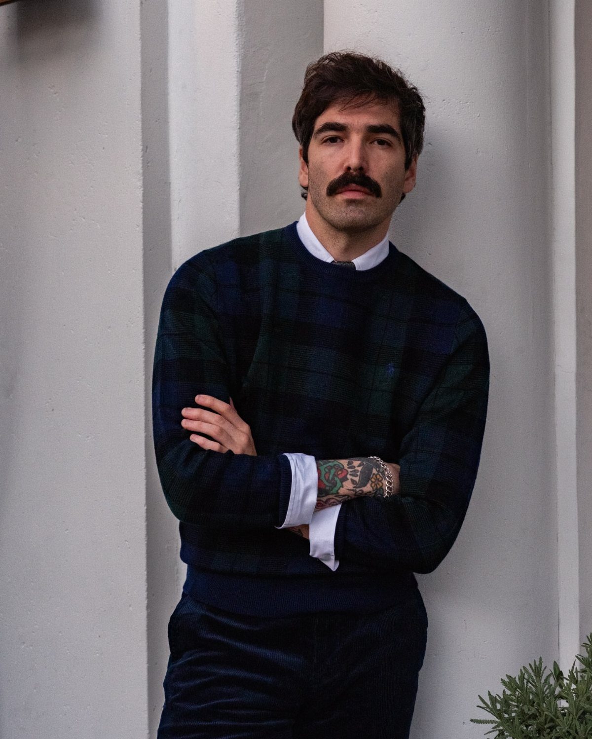 Man with mustache wearing a plaid sweater and collared shirt against a white wall.