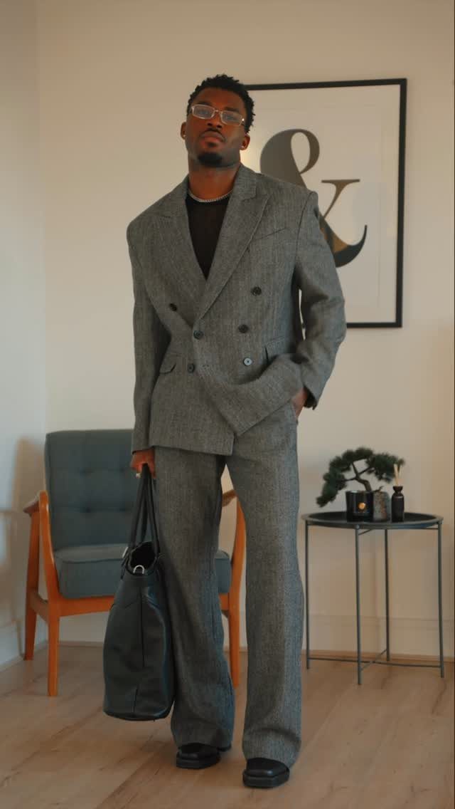 Man in a grey double-breasted tailored suit holding a black leather bag indoors.