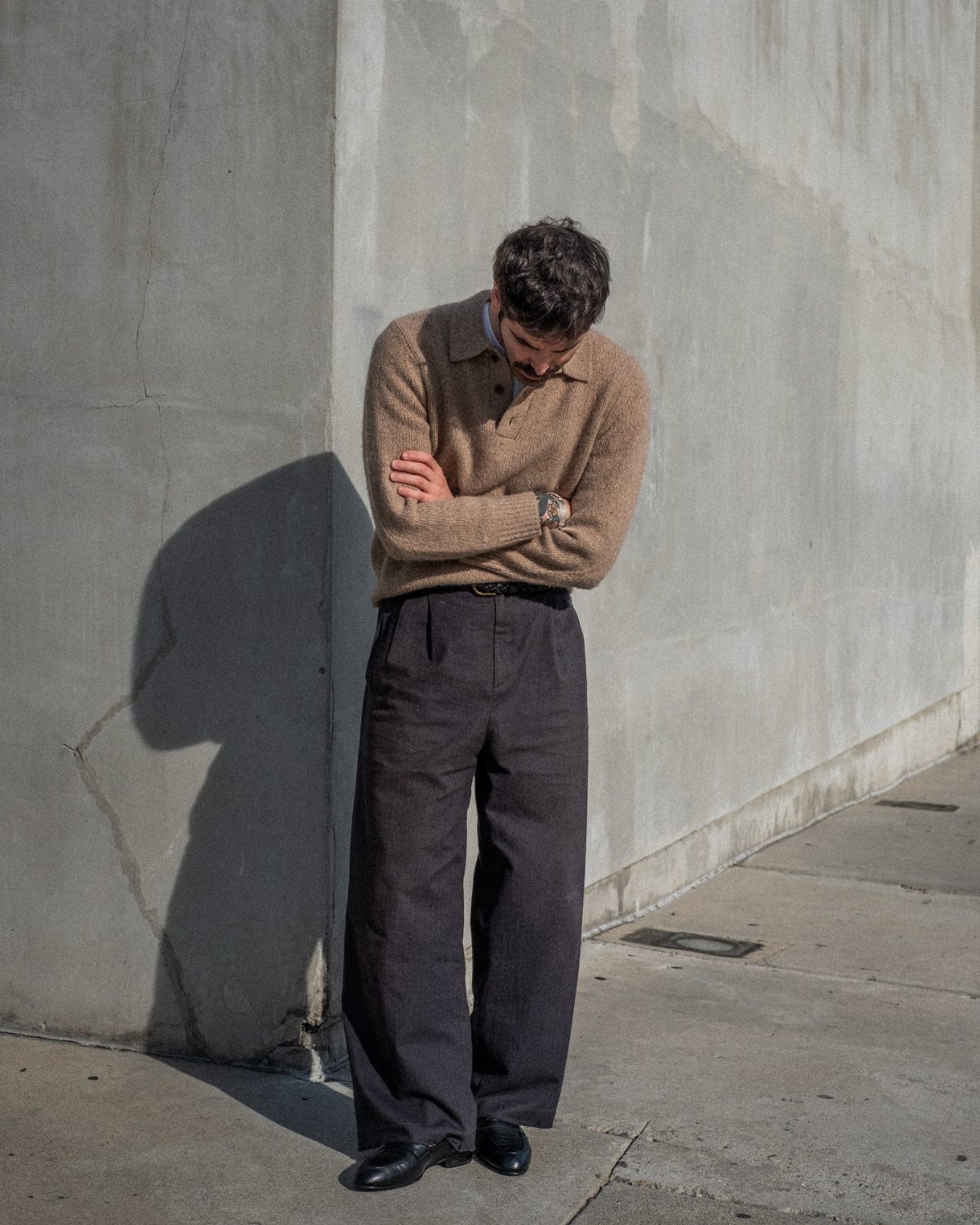 Man in brown knit polo and wide-leg grey trousers standing by a concrete wall.