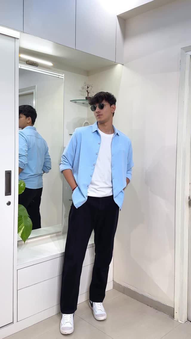 Man wearing an open light blue shirt over a white t-shirt and black trousers.