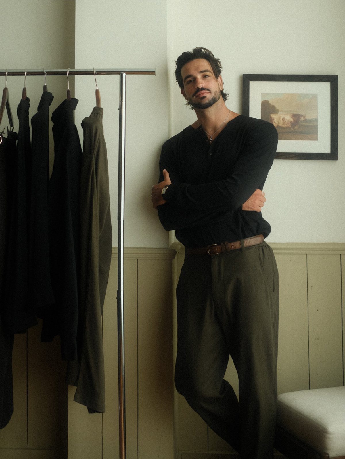 Man in black shirt and olive trousers standing next to a clothing rack.